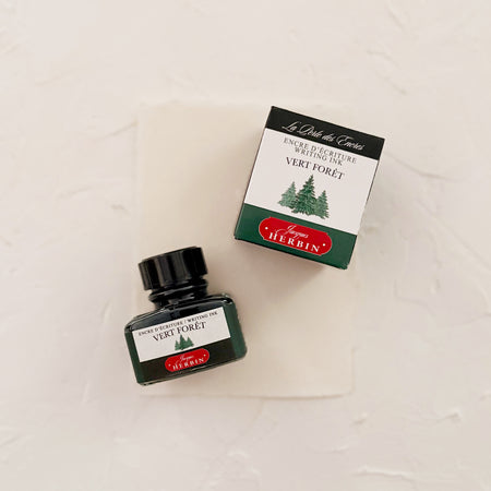 Glass square bottle with black cap. White label with black text saying, “Vert Foret” and white text saying, “HERBIN”. Green stripe on each side of bottle. bottle next to box with the same label. both on textured background
