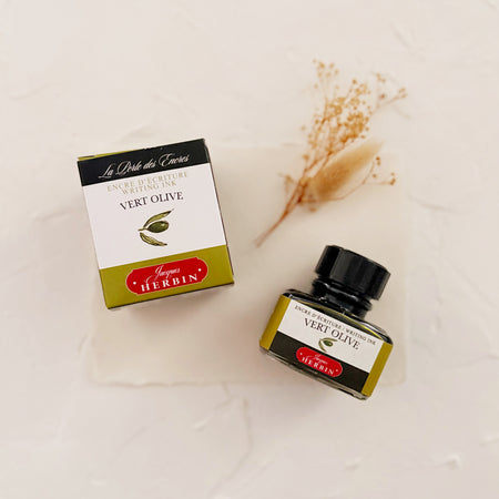 Glass square bottle with black cap. White label with black text saying, “Vert Olive” and white text saying, “HERBIN”. Olive green stripe on each side of bottle. bottle is next to box with the same label, on textured background and cream dried florals