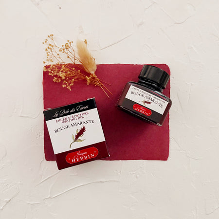 Glass square bottle with black cap. White label with black text saying, “Rouge Amarante” and white text saying, “HERBIN”. Burgandy stripe on each side of bottle. bottle next to box with same label on textured maroon background