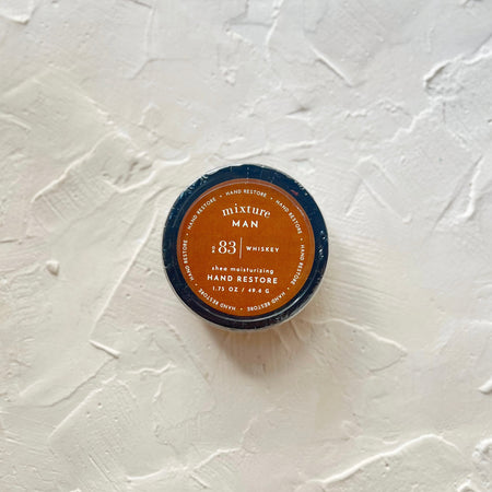 Black circle container with orange label with white text saying, “Mixture Man No 83 Whiskey Moisturizing Hand Restore”.
