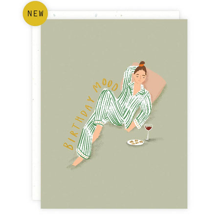 Olive card with gold foil text saying, “Birthday Mood.” Image of a woman wearing green striped pajamas lounging on a pillow with a wine glass and a cheese plate. A white envelope is included.