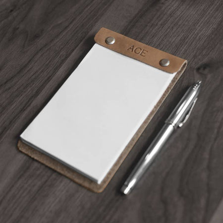 White notepad bound in brown leather case with two brass screws on either side. 