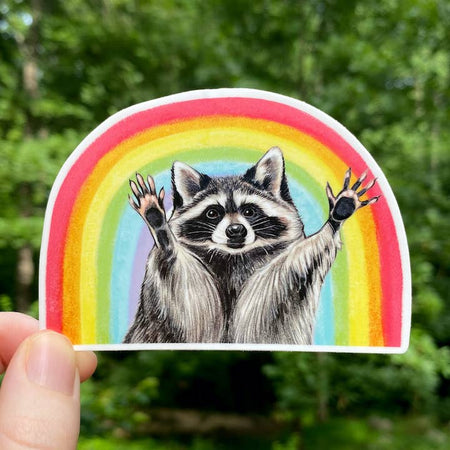 Sticker in the image of a raccoon raising his hands up with a rainbow in the background.