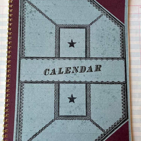 Maroon cover with light blue geometric shape design in center with dark blue text saying, “Calendar”. A gold coil binding on left side.