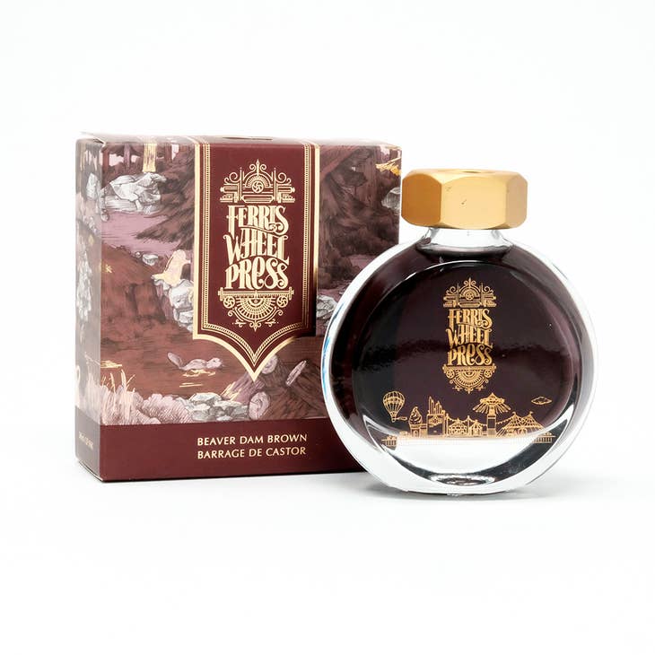 Round glass bottle with gold cover and gold text saying, "Ferris Wheel Press" with images of a carnival on front of bottle. Ink is wine colored. Packaged in square wine purple box with images of a beaver dam.