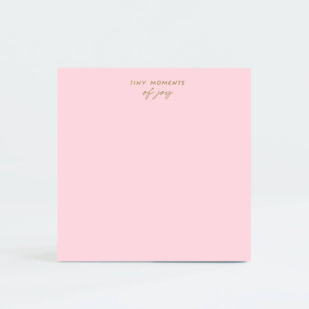 Pink square notepad with gold foil text saying, “Tiny Moments of Joy” in top center.