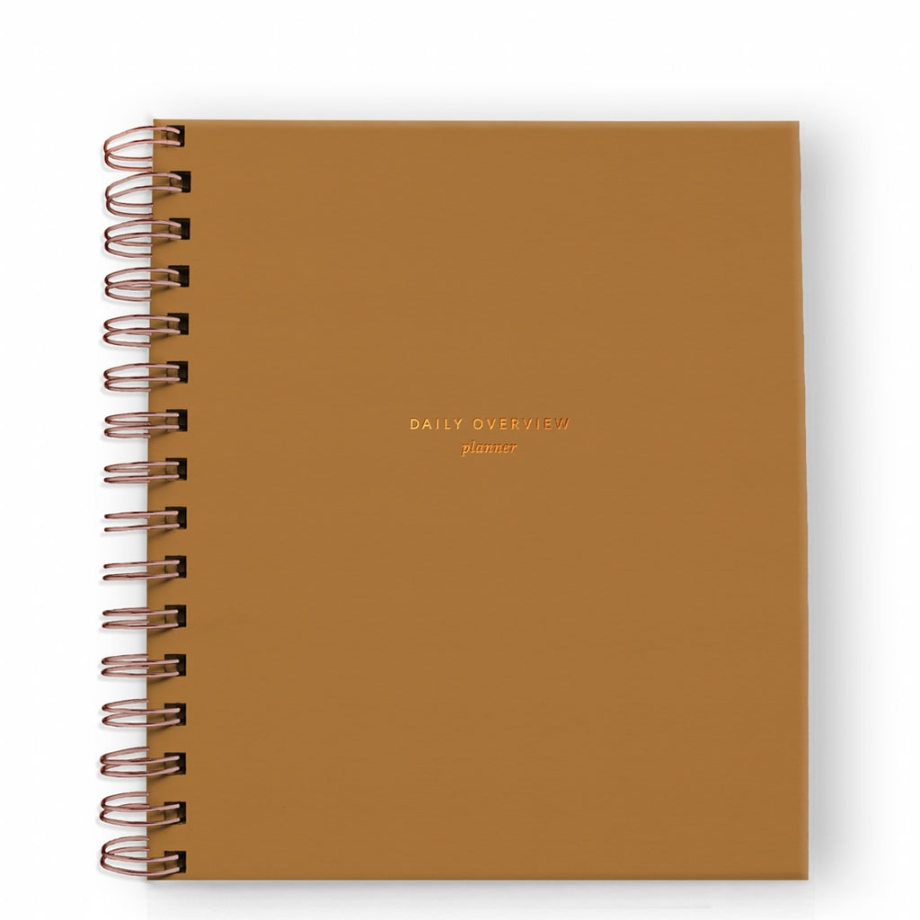 Notebook with mustard orange cover with gold foil text saying, “Daily Overview Planner”. Gold coil binding on left side.
