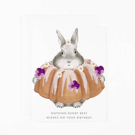 White card with image of a gray bunny holding a bundt cake with purple flowers and white icing. Gray text saying, “Nothing Bundt Best Wishes on Your Birthday”. A white envelope is included.