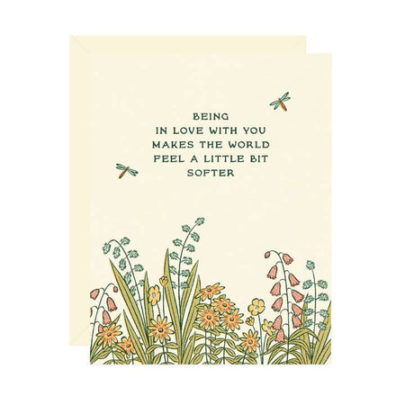 White card with images of wildflowers on the bottom and dragonflies on the top. Blue text saying, “Being in Love with You Makes the World Feel A Little Bit Softer”. A white envelope is included.