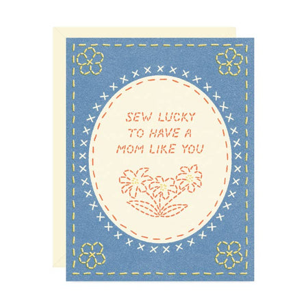 Blue card with images of a hand sewn picture with flowers in the corner and white oval in center with sewn text saying, “Sew Lucky to Have a Mom Like You”. A white envelope is included.