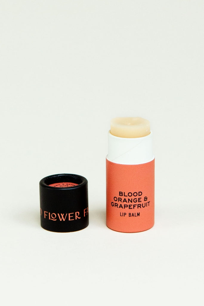 Orange tube with black cover. Cover has orange text saying, “Good Flower Farm”. Tube has black text saying, “Blood Orange & Grapefruit Lip Balm”.  Balm is an ivory color.