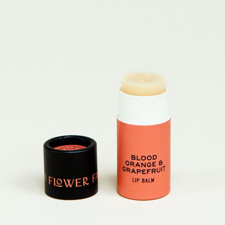 Orange tube with black cover. Cover has orange text saying, “Good Flower Farm”. Tube has black text saying, “Blood Orange & Grapefruit Lip Balm”.  Balm is an ivory color.