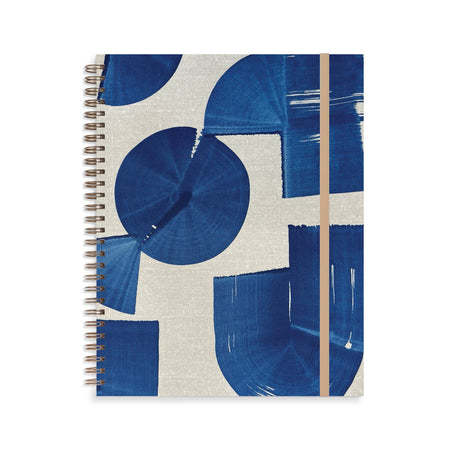 Notebook with ivory cover with indigo blue abstract shapes. Metal coil binding on left side. Tan elastic over right side.