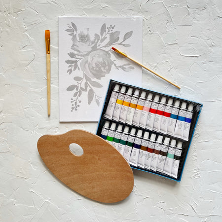 Painting kit with white canvas with gray floral design; brown brush, wooden brown palette, and black case with various colored paint tubes.