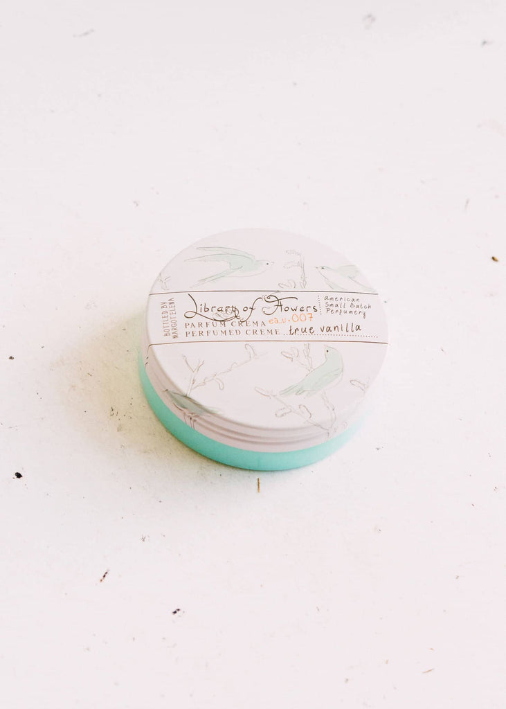 Circular package with white lid with muted images of bluebirds on a branch. Black text saying, “Margot Elena Library of Flowers Parfum Crema True Vanilla”.