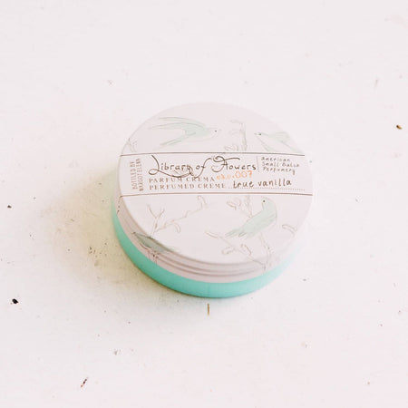 Circular package with white lid with muted images of bluebirds on a branch. Black text saying, “Margot Elena Library of Flowers Parfum Crema True Vanilla”.