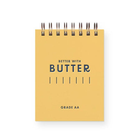 Yellow notebook with blue text saying, “Better with Butter”. Made in the image of a stick of butter. Gold coil binding across top.