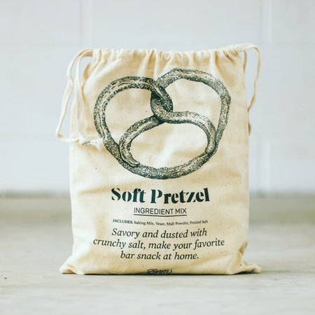 Ivory drawstring bag with black text saying, “Soft Pretzel: Savory and dusted with crunchy salt, make your favorite bar snack at home.” Image of a large twisted pretzel.