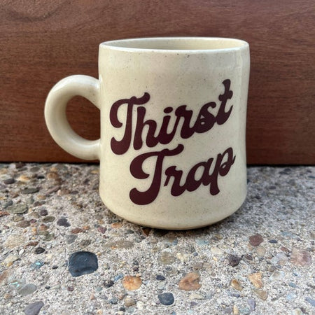 Ivory mug with tiny brown speckles and brown text saying, “Thirst Trap”.