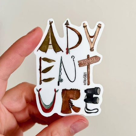 Hand holding a sticker with 'ADVENTURE' text and travel-themed illustrations.