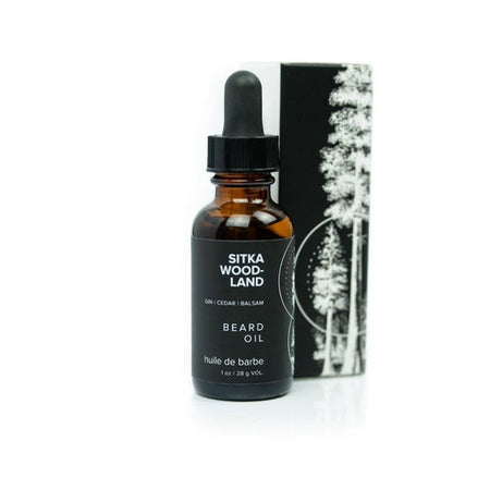 Brown bottle with dropper top and black label with white text saying, “Sitka Woodland Beard Oil”. Image of a white and black pine trees on side of label.