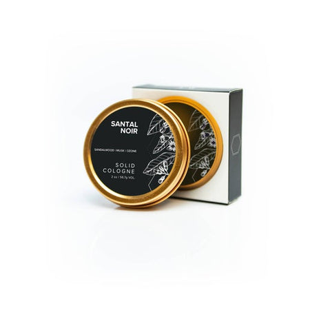 Round gold tin with black circle label with white text saying, “Santal Noir Solid Cologne”. Image of a black and white leaves on side of label.