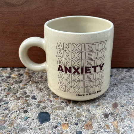 Ivory mug with tiny brown speckles and brown text saying, “Anxiety Have A Nice Day”.