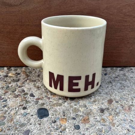 Ivory mug with tiny brown speckles and brown text saying, “Meh”.