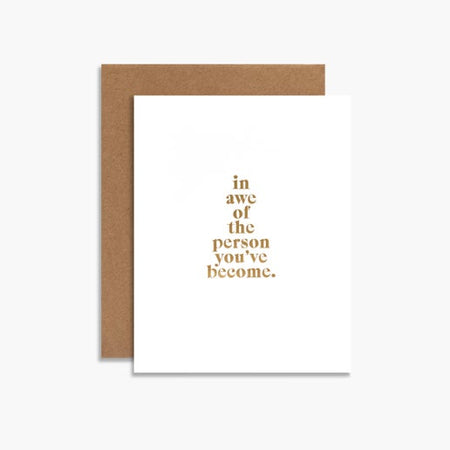 White card with gold foil text saying, “In awe of the person you’ve become”. A brown kraft envelope is included.