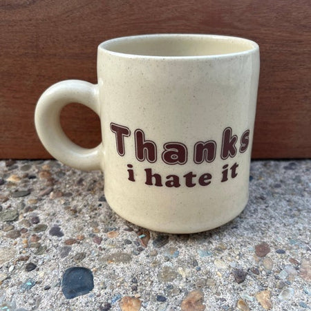 Ivory mug with tiny brown speckles and brown text saying, “Thanks I Hate It”.
