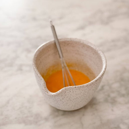 Small white speckled ceramic bowl with pouring spout and a small metal whisk.