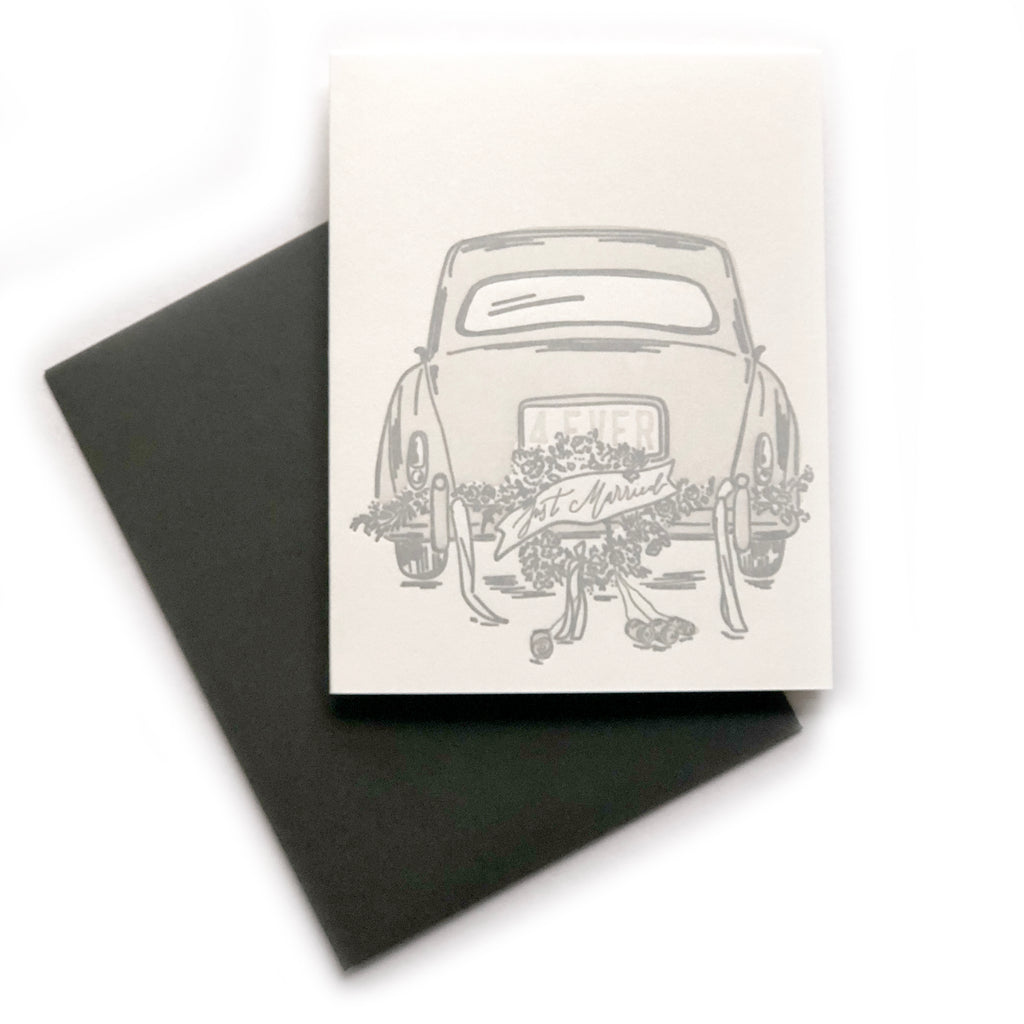 White card with image of the back of a silver card with flowers and ribbons attached to the tailgate with a banner saying, “Just Married”. A black envelope is included.