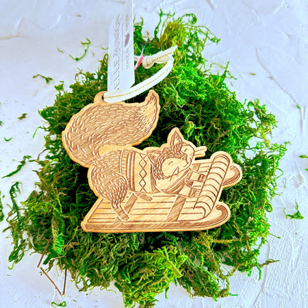 Wooden carved ornament of a fox wearing a winter sweater and riding on a sled.