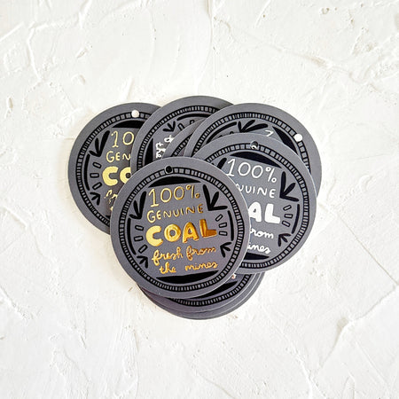 Circular gray gift tags with gold text saying, “100% Genuine Coal Fresh From the Mines”. Images of black embellishments to make the appearance of a coin.