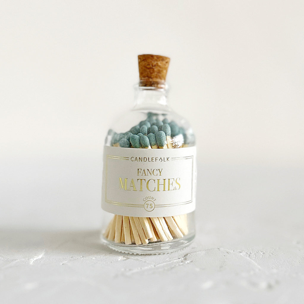 Small glass bottle with cork lid and white label with gold text saying, “Candlefolk Fancy Matches”. Filled with wooden matches with slate green tops.