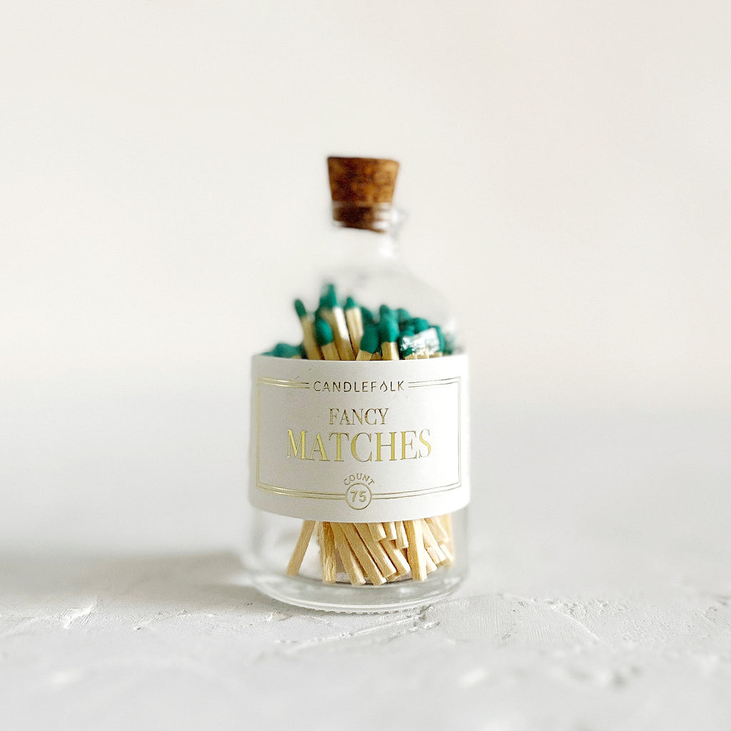 Small glass bottle with cork lid and white label with gold text saying, “Candlefolk Fancy Matches”. Filled with wooden matches with green tops.