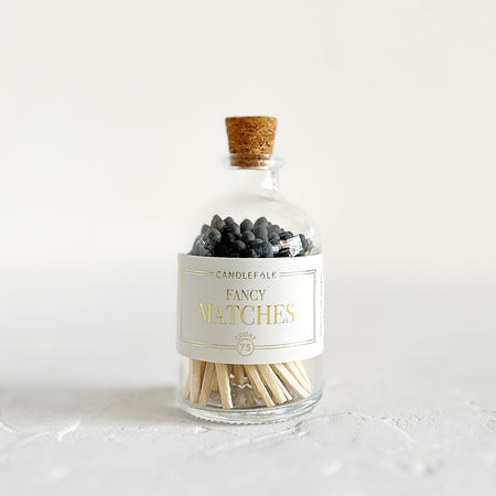 Small glass bottle with cork lid and white label with gold text saying, “Candlefolk Fancy Matches”. Filled with wooden matches with black tops.