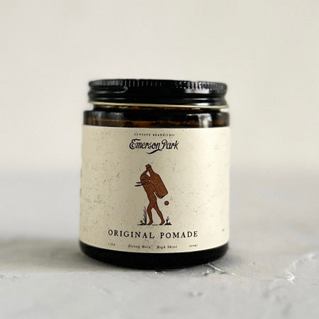 Brown jar with a black cover and a white label saying, “Emerson Park Original Pomade”. Image of a man carrying a large wine bottle on his back. 
