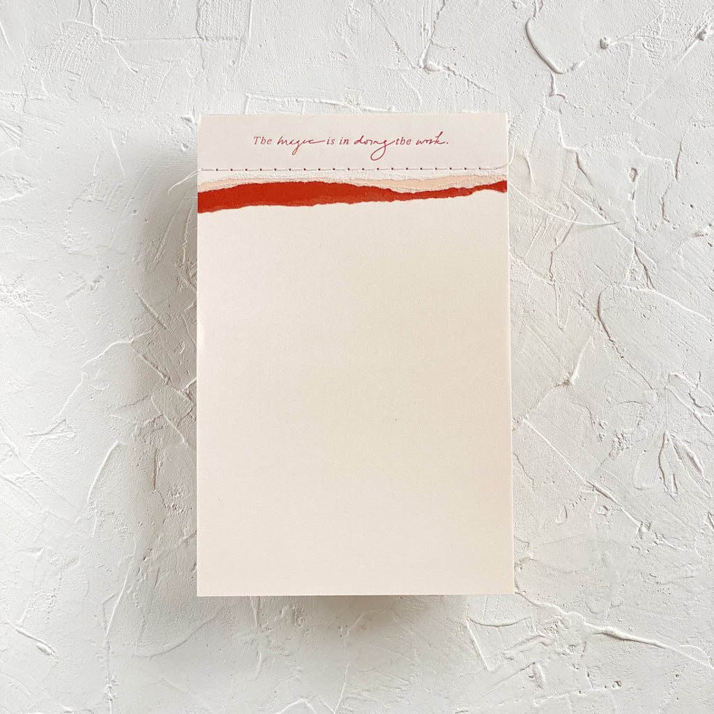 Ivory notepad with pink and red torn edging across the top with red text saying, “The Magic is in Doing the Work”. 
