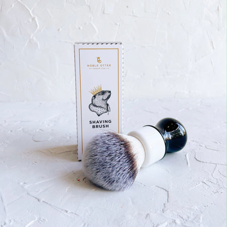 Packaged in a white tall rectangle box with gold and blue text saying, “Noble Otter Soap Co. Shaving Brush”. Actual brush has black and white handle with white, black and blue bristles.