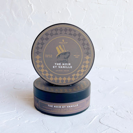 Circular blue packaging with gold and white text saying, “Noble Otter Soap Co. The Noir Et Vanille Shaving Soap”. Images of gold checkerboard around edge and an otter wearing a gold top hat in center.