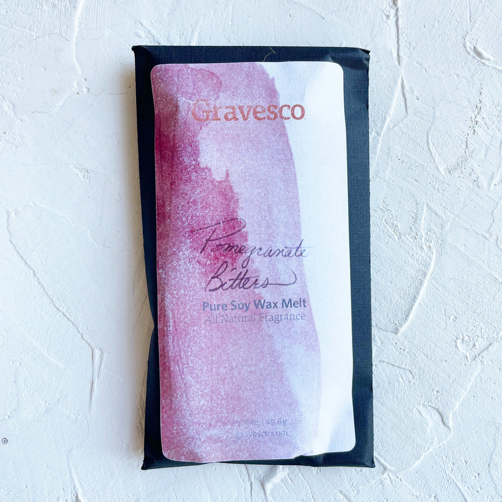 Packaged in a black rectangle pouch with white and red label with black and red text saying, “Gravesco Pomegranate Bitters Wax Melt”.