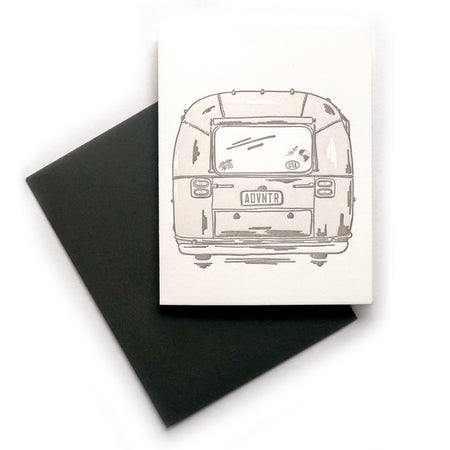 Ivory card with image of the backside of a silver vintage camper with license plate saying, “ADVNTR” and images of a globe, paper airplane, red heart, USA and Adventure Awaits on the back window. A black envelope is included.