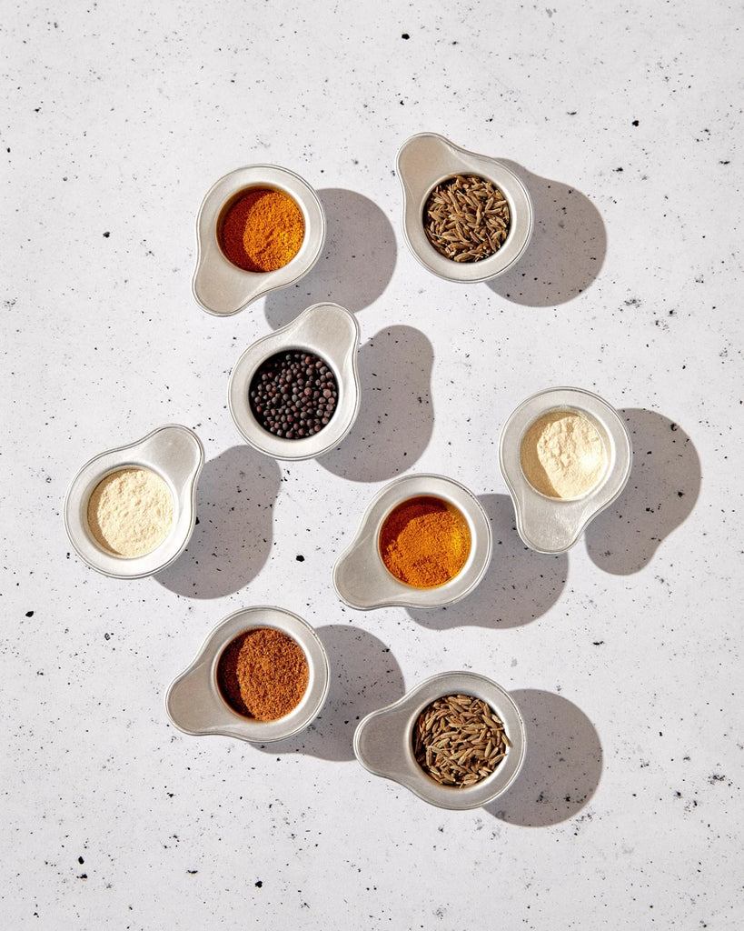 Images of several spices in browns, ivory, black and greens packaged in round white cups.