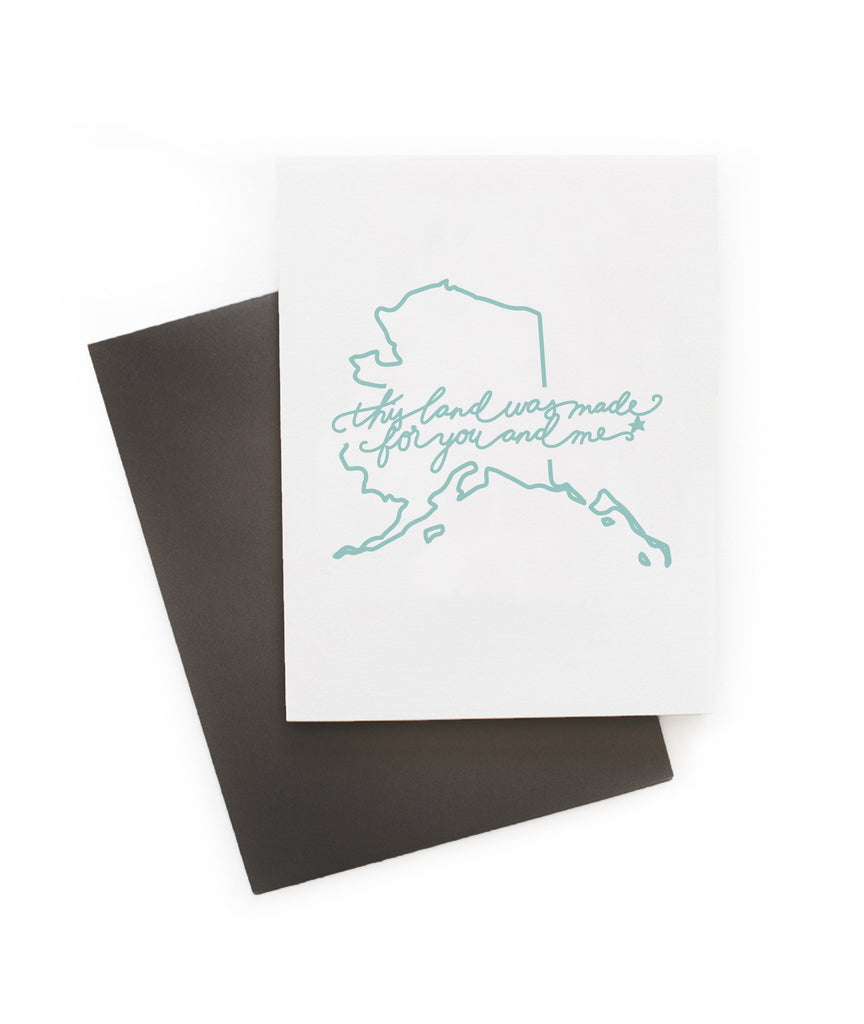 White card with teal text saying, “This Land is Made for You and Me”. Teal outline image of the state of Alaska. A gray envelope is included.