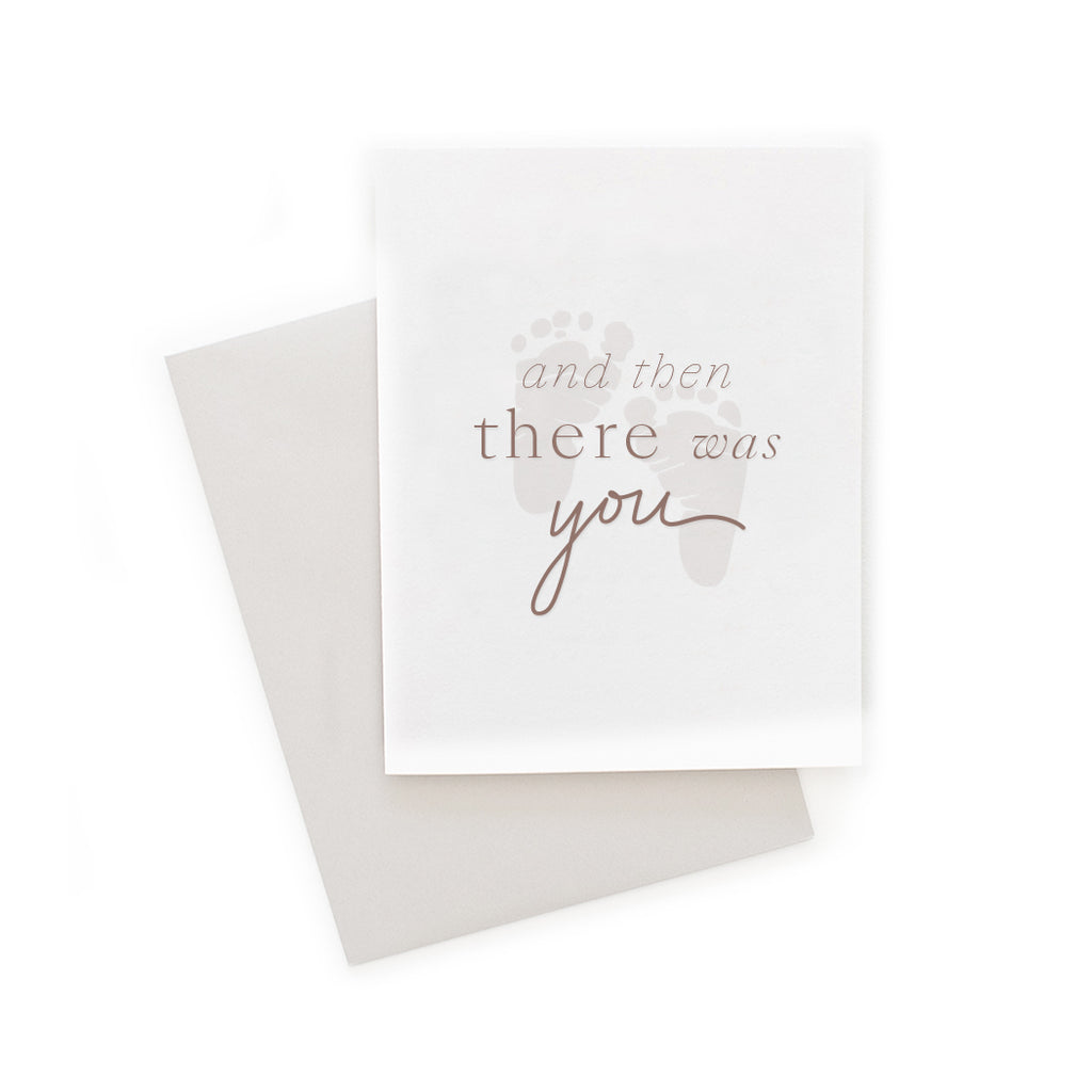 White card with pink text saying, “And Then There Was You”. Images of gray baby’s footprint in middle of card. A gray envelope is included.