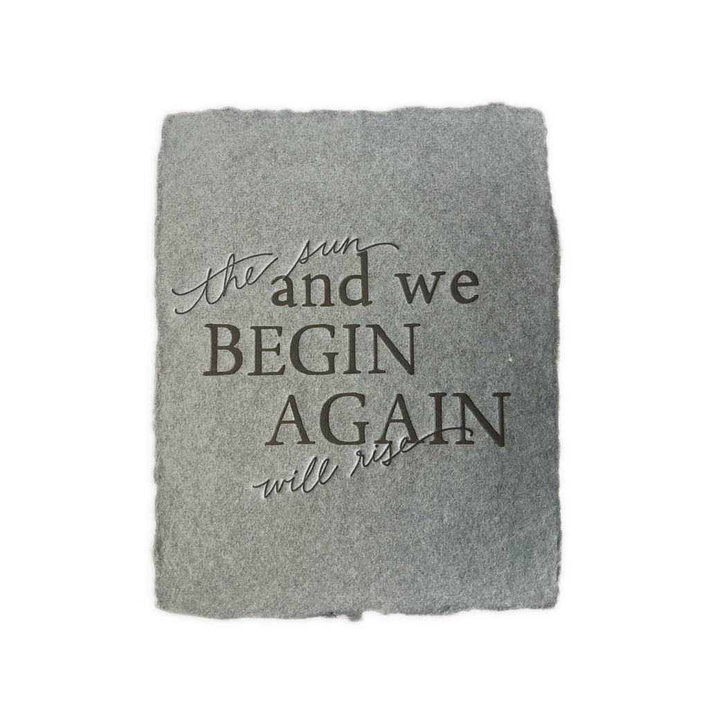 Gray card with texture to look like cement with black text saying, “The Sun Will Rise and We Begin Again”. An envelope is included.