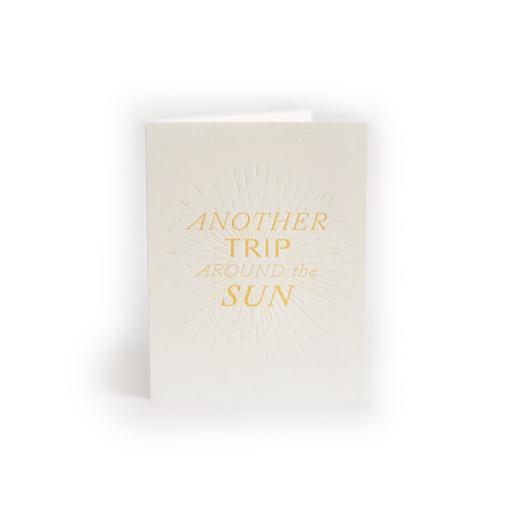 Ivory card with yellow text saying, “Another Trip Around the Sun”. Images of embossed sun rays shooting around the text. An envelope is included.