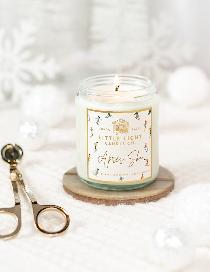 Glass jar with gold lid and white label with gold foil text saying, “Little Light Candle Co. Apres Ski”. Images of various skiers on label.