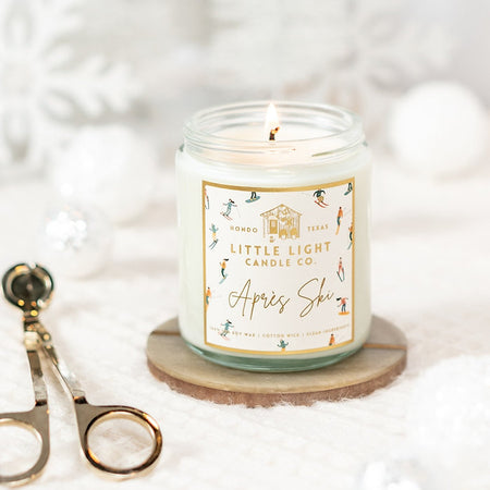 Glass jar with gold lid and white label with gold foil text saying, “Little Light Candle Co. Apres Ski”. Images of various skiers on label.